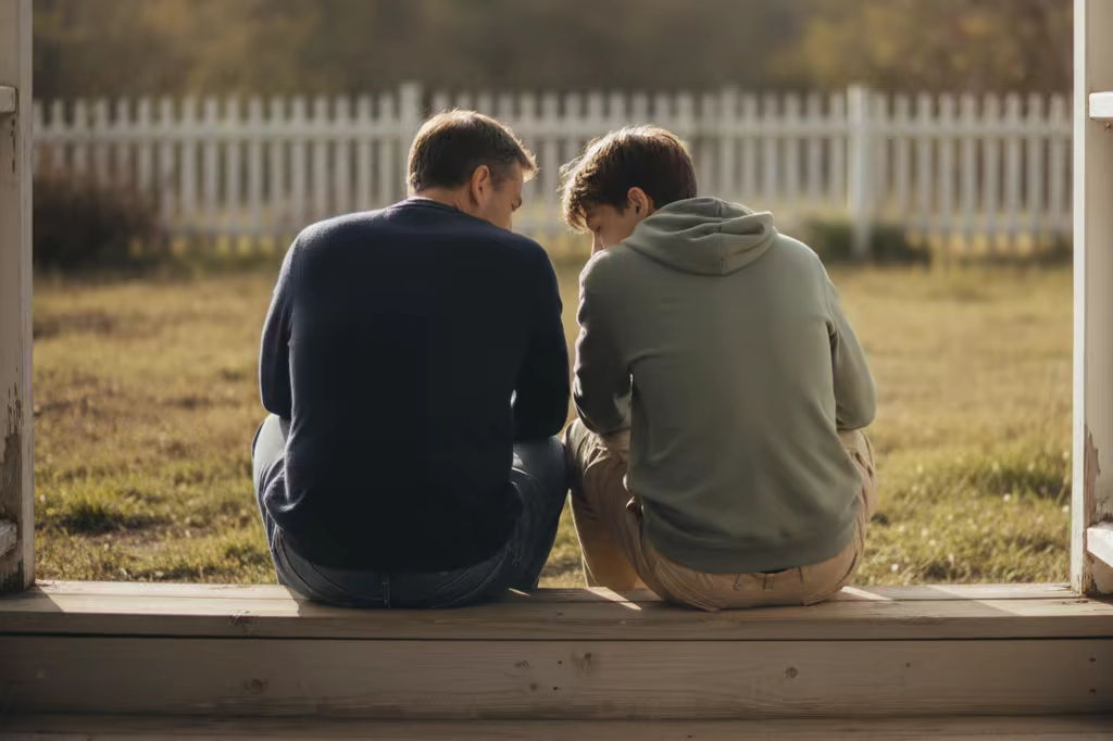 A parent and teenager sitting on a porch step in conversation, seen from a distance