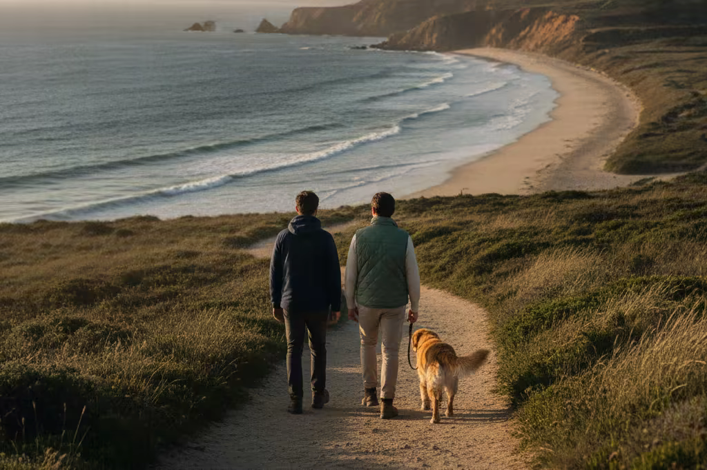 Two people walking a dog on a coastal path in late afternoon light, seen from behind