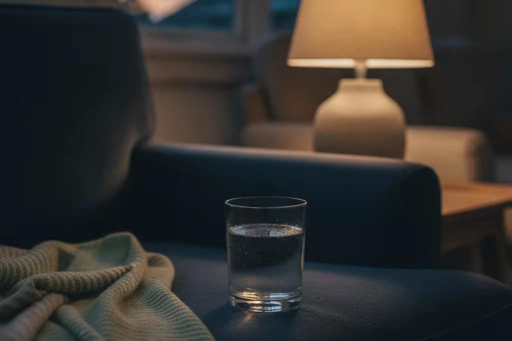 A glass of water and a blanket on a couch beside a dim lamp in a quiet living room